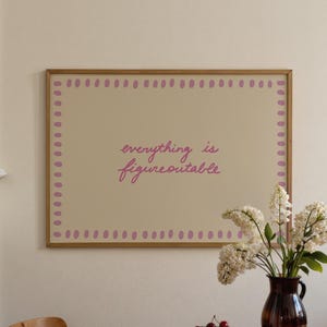 May include: A framed print with the words "everything is figureoutable" in a cursive font. The print has a light beige background with a border of small purple dots. A vase of white flowers sits on a wooden table.