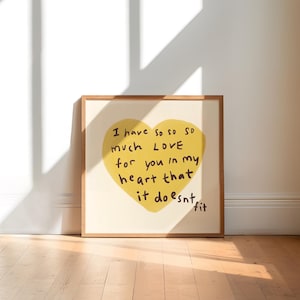 May include: A square framed print featuring a large yellow heart on a cream background. The heart contains the handwritten black text: I have so so so much LOVE for you in my heart that it doesnt fit. The frame is made of light-coloured wood.