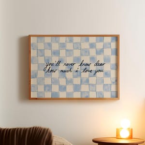 Può includere: Opera d'arte con cornice in legno con un motivo a scacchi blu e bianco. L'opera d'arte presenta il testo scritto a mano: "you'll never know dear how much I love you." L'opera d'arte è esposta sopra un letto con una coperta marrone.