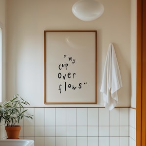 Könnte beinhalten: Badezimmerinterieur mit weißer Badewanne, Waschbecken und einem gerahmten Druck mit dem Text "my cup overflows". Der Raum verfügt über weiß geflieste Wände, einen Holzwaschtisch und eine Topfpflanze. Ein weißes Handtuch hängt an der Wand.