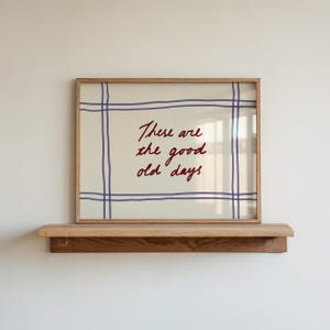 Pode incluir: Uma impressão emoldurada com o texto "These are the good old days" em escrita vermelha. A impressão tem um fundo creme com um padrão de grade azul e é exibida em uma prateleira de madeira.