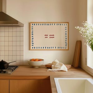 May include: A framed artwork with the text "mi casa su casa" in red script. The artwork is surrounded by blue dots and has a wooden frame. The kitchen scene includes a bowl of oranges, a cutting board, and a gas stove.