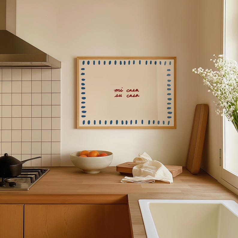 May include: A framed artwork with the text "mi casa su casa" in red script. The artwork is surrounded by blue dots and has a wooden frame. The kitchen scene includes a bowl of oranges, a cutting board, and a gas stove.