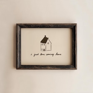Puede incluir: Una impresión artística enmarcada con una ilustración en blanco y negro de una casa y la frase manuscrita, "i just love coming home". El marco es marrón oscuro con un acabado envejecido. La obra de arte es una representación sencilla y encantadora de una casa.