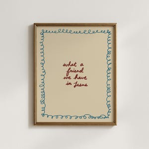 Puede incluir: Obra de arte enmarcada con el texto bordado "what a friend we have in Jesus" en escritura burdeos. La obra está enmarcada en un marco dorado, con un borde bordado azul. El fondo es de color beige claro.