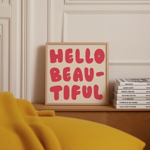 Peut inclure: Une affiche encadrée avec les mots "HELLO BEAUTIFUL" en lettres rouges, de style bulle, sur un fond crème. Le cadre est marron clair. Une pile de livres est à droite de l'affiche.