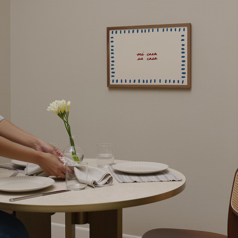 May include: A dining table set for a meal, featuring a round table with a beige top and a gold-colored base. The table is set with white plates, glasses, and folded napkins. A framed artwork with the text "mi casa su casa" hangs on the wall.