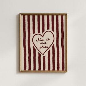 May include: A framed wall art print featuring a cream background with vertical dark red textured stripes. In the centre, a dark red outlined heart contains the handwritten-style text "this is our place" in dark red ink on a cream background.