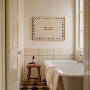 May include: A framed artwork with the text "mi casa su casa" in red and a blue dotted border hangs above a white bathtub. The bathroom features a wooden stool, a bottle, and a black and white checkered floor.