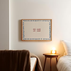 May include: A framed artwork with the text "mi casa su casa" in red and blue. The artwork is surrounded by a border of blue dashes and has a natural wood frame. The artwork is hanging on a white wall.