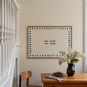 May include: A framed artwork with the text "mi casa su casa" in red script. The artwork has a cream-colored background with a border of blue dashes. A wooden table holds a vase of white flowers and a plate of cherries.