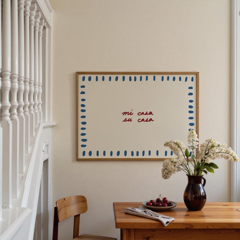 May include: A framed artwork with the text "mi casa su casa" in red script. The artwork has a cream-colored background with a border of blue dashes. A wooden table holds a vase of white flowers and a plate of cherries.