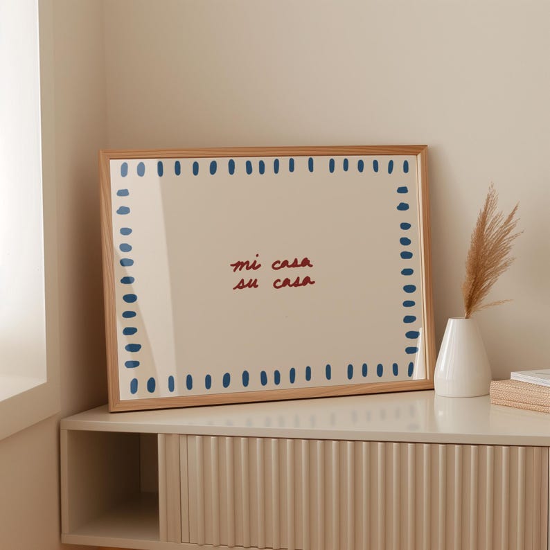 May include: A framed art print with the text "mi casa su casa" in red and blue. The print has a light beige background with a border of blue dots. The frame is made of light brown wood.