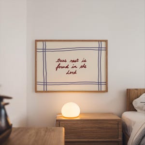 Pode incluir: Uma obra de arte emoldurada com o texto "true rest is found in the Lord" em escrita vermelha. A obra de arte tem um fundo creme com linhas de grade azuis e uma moldura de madeira. Uma pequena lâmpada redonda e brilhante está em uma mesa de cabeceira de madeira.