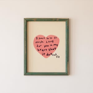 May include: A framed art print with a green wooden frame. The artwork features a pink heart with the handwritten text "I have so so much LOVE for you in my heart that it doesn't fit."