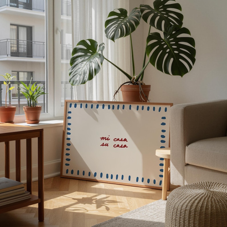May include: A framed artwork with the text "mi casa su casa" in red script. The frame is wood with blue dotted accents. A large potted Monstera plant sits atop the frame, with a neutral-toned sofa and a small side table in the background.