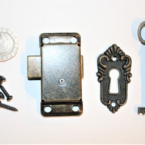 May include: An assortment of antique-style hardware components, including a metal lock, key, keyhole cover, and screws. The hardware has a bronze finish. A British 20 pence coin is also present.