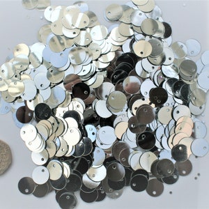 May include: A collection of shiny, silver sequins scattered on a white surface. Each sequin is round with a small hole at the top. A coin is included for size reference. These are ideal for crafting and embellishments.