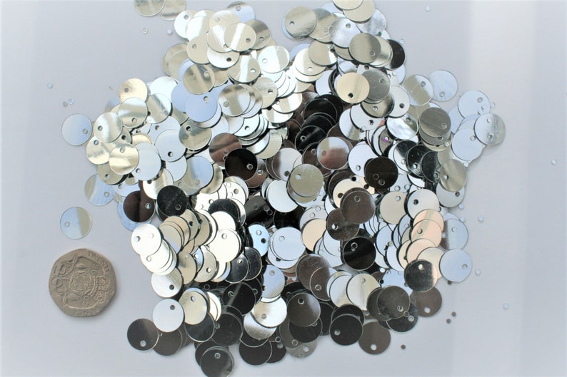 May include: A collection of shiny, silver sequins scattered on a white surface. Each sequin is round with a small hole at the top. A coin is included for size reference. These are ideal for crafting and embellishments.
