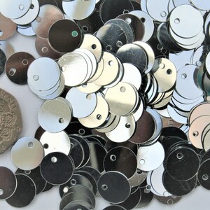 May include: A collection of shiny, silver-colored sequins with a small hole in the center. A British twenty pence coin is included for size comparison. The sequins are round and reflect light.