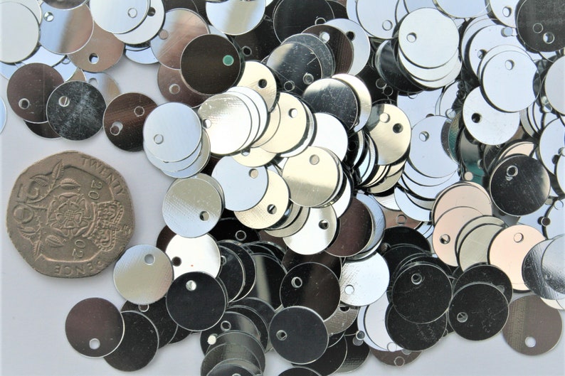 May include: A collection of shiny, silver-colored sequins with a small hole in the center. A British twenty pence coin is included for size comparison. The sequins are round and reflect light.