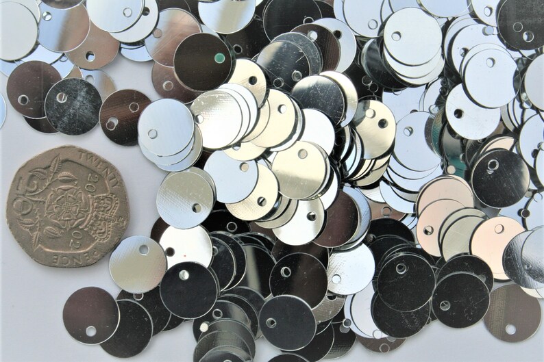 May include: A collection of shiny, silver-colored sequins with a small hole in the center. The sequins are round and scattered across a white surface. A twenty pence coin is also present for size comparison.