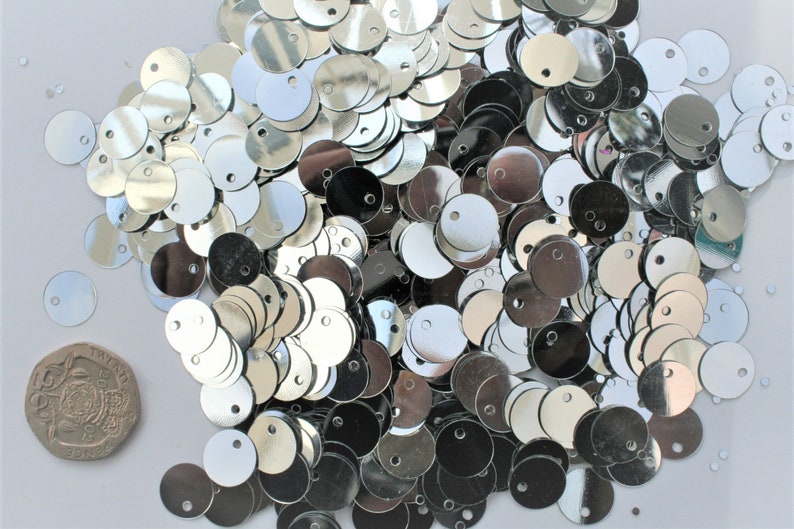 May include: A pile of shiny, silver sequins with a small hole in the center. The sequins are round and reflective, scattered across a white surface. A twenty pence coin is visible for size comparison.