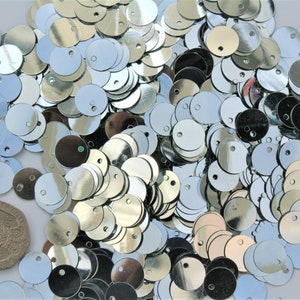 May include: A collection of shiny, silver sequins scattered across a surface. Each sequin is round with a small hole near the top. A British twenty pence coin is also visible, providing a size reference. The sequins reflect light, creating a sparkling effect.