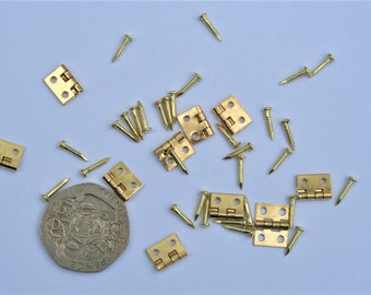 May include: An assortment of small, gold-colored hinges and nails scattered on a light blue surface. The hinges are rectangular with two leaves and a central hinge. The nails are small and pointed. A coin is also present.