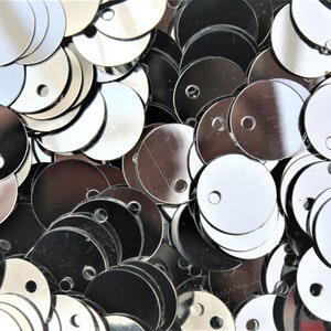 May include: Close-up of a pile of shiny, silver sequins. The sequins are round with a small hole near the top for stringing. Some sequins are reflective, while others appear darker. The sequins are scattered, creating a textured surface.