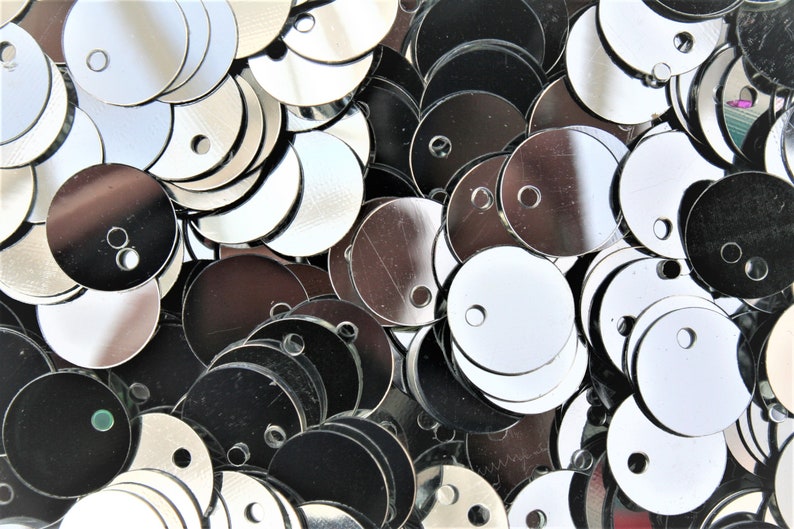 May include: Close-up of a pile of shiny, silver sequins. The sequins are round with a small hole near the top for stringing. Some sequins are reflective, while others appear darker. The sequins are scattered, creating a textured surface.