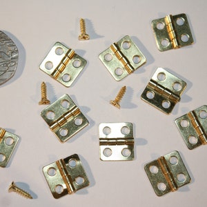 Puede incluir: Un conjunto de pequeñas bisagras y tornillos dorados, probablemente para proyectos en miniatura o manualidades. Las bisagras son cuadradas con esquinas redondeadas y cuatro agujeros para tornillos. Se incluye una moneda como referencia de tamaño.