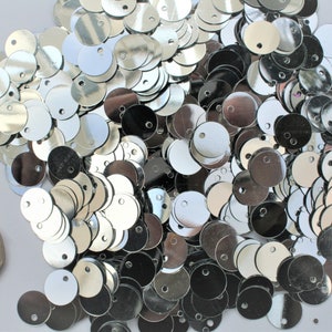 May include: A pile of shiny, silver sequins, each with a small hole for sewing. The sequins are round and reflective, scattered across a surface. A coin is included for size reference.