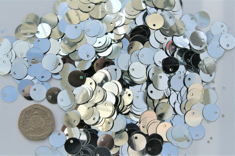 May include: A collection of shiny, silver sequins scattered across a surface. Each sequin is round with a small hole near the top. A British twenty pence coin is also visible, providing a size reference. The sequins reflect light, creating a sparkling effect.