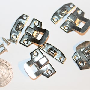 May include: A collection of silver-colored metal latches, screws, and a coin. The latches have a rectangular design with a hinged closure. The screws are small and pointed. The coin is a twenty pence piece.