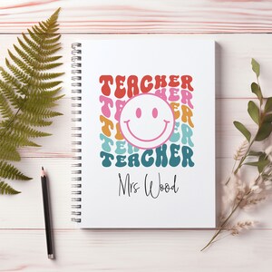 May include: White spiral-bound notebook featuring the word "TEACHER" in a retro font, a smiley face, and the name "Mrs Wood". A pencil and fern are on the left, with greenery on the right. The notebook is on a light wood surface.