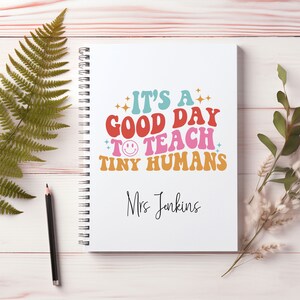 May include: A white spiral notebook with the text "It's a good day to teach tiny humans" in colourful retro font. The name "Mrs. Jenkins" is written below the text.