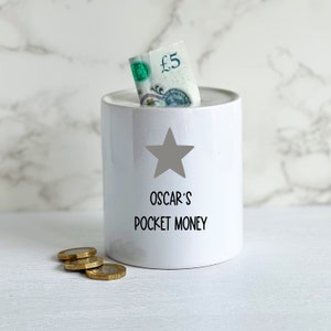 May include: White ceramic money box with a grey star and the text "Oscar's Pocket Money". A five pound note and some coins are in the box.