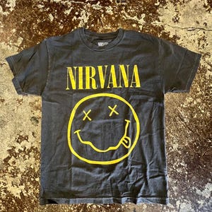 Puede incluir: Una camiseta de manga corta en color gris carbón que presenta el logotipo de Nirvana en amarillo y un gráfico de cara sonriente amarilla con ojos en forma de X y la lengua fuera. La prenda tiene cuello redondo y un corte estándar con estampado.