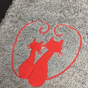 May include: Gray towel with a red embroidered design of two cats inside a heart-shaped outline. The cats are stylized with long bodies and pointed ears. The towel has a looped texture.