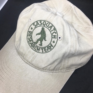 May include: A beige baseball cap with a green embroidered circle logo that says "Sasquatch Research Team" and features a green silhouette of a sasquatch walking.