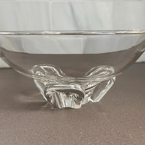 May include: A clear glass bowl with a wide, shallow design. The bowl has a rounded rim and sits on three decorative, sculpted feet. The glass is transparent, allowing light to pass through, and the bowl appears to be empty.