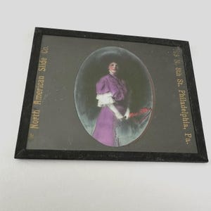 May include: Antique framed slide featuring a woman in a purple dress with white lace sleeves, set against a dark background. The frame has text that reads "North American Slide Co." and "133 N. 8th St. Philadelphia, Pa."