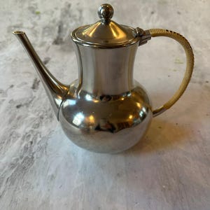 May include: A silver-colored teapot with a rounded body, a long spout, and a lid with a small ball on top. The handle is wrapped with a light-colored woven material. The teapot is likely made of stainless steel.