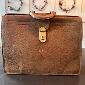 May include: A vintage brown leather briefcase with a textured surface. The briefcase has a handle, a metal clasp, and the initials "E.S.L." embossed on the front. The briefcase is rectangular and appears to be well-worn.