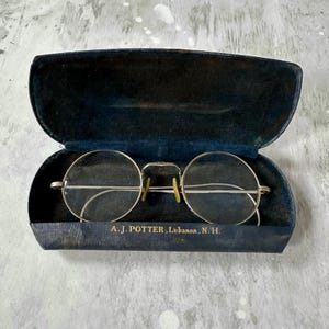 May include: Vintage round eyeglasses with a silver frame and clear lenses, resting inside a dark blue case. The case is open, revealing the glasses and the text "A.J. POTTER, Lebanon, N.H." on the inside.