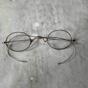 May include: Antique round eyeglasses with a thin, bronze-colored metal frame. The lenses are clear, and the earpieces curve downward. The glasses are set against a mottled gray background.