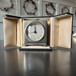 May include: A silver-toned square travel clock with a white face and black numerals. The clock is displayed inside a black case with beige lining. The clock has a classic design and is likely vintage.