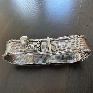 May include: A silver-toned mesh bracelet with a decorative ball and ornate clasp. The bracelet is wide and has a woven texture. The clasp is a simple bar that secures the bracelet. The bracelet is lying on a dark surface.