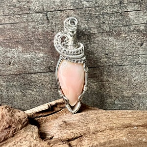 Peut inclure: Pendentif artisanal avec une pierre précieuse rose pâle en forme de goutte d'eau, sertie dans un fil d'argent complexe. Le fil forme une bélière décorative et enveloppe la pierre, créant un bijou unique et élégant.